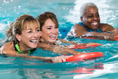 Three women joyfully participate in an adult learn to swim class, holding red kickboards as they move through the water.