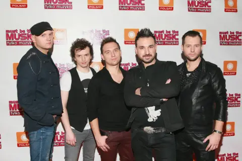 Pięciu mężczyzn w strojach scenicznych pozuje do zdjęcia na tle ścianki z logo "Must Be The Music".