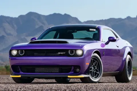 En lila amerikansk sportbil, en Dodge Challenger SRT, står parkerad på en grusväg med berg i bakgrunden.