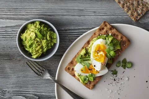 Delicioso sandwich de aguacate y huevo sobre pan integral, con un tazón de aguacate machacado y un tenedor al lado.