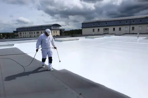 Un ouvrier applique une peinture réflective toiture blanche sur un toit plat. Le ciel est couvert de nuages sombres.
