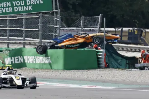 Wypadek bolidu F1 na torze Monza. Pomarańczowo-niebieski bolid leży na bandzie, ratownicy udzielają pomocy.