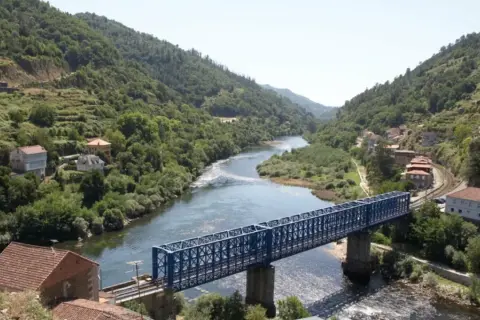 Un puente azul cruza un río serpenteante rodeado de colinas verdes y casas. El paisaje evoca la tranquilidad de los peares en un día soleado.