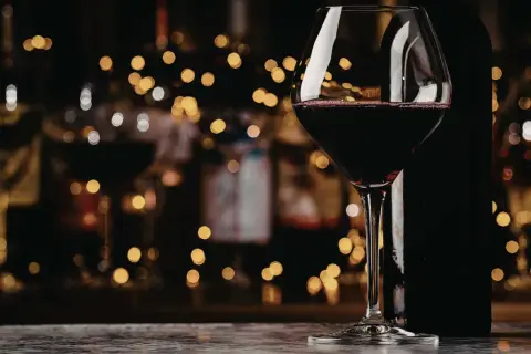 Szklanka z czerwonym winem, obok butelka. W tle migoczące światła, jakby wieczór w winiarni. To wino bycza moc.