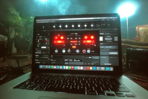 Laptop screen displays a vocal chorus plugin interface, with glowing red knobs and sliders, set in a dimly lit room with plants and furniture.