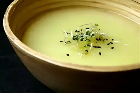Un bol en bois contient un velouté d'asperges blanches, garni de pousses vertes et de graines noires.