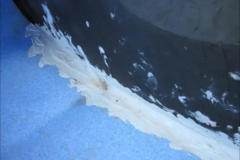 Application d'un mastic blanc pour coller du polyéthylène sur une surface bleue texturée, avec des éclaboussures sur le fond sombre.
