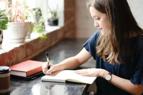 Une jeune femme écrit dans un carnet, peut-être le début de sa propre biographie. À côté, un livre rouge et une tasse de café.