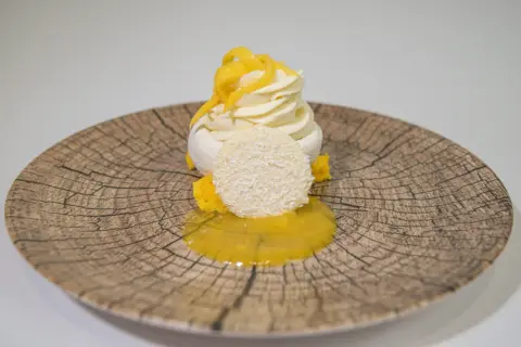 Deliciosa pavlova de mango con merengue, crema batida, coco rallado y salsa de mango sobre un plato rústico.