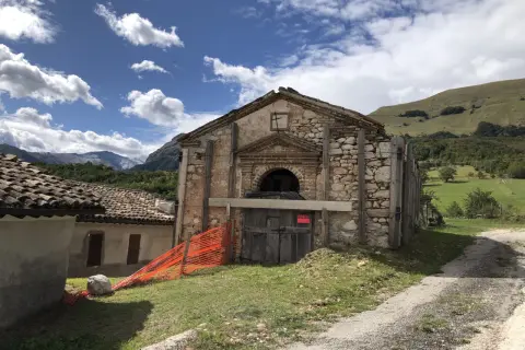 Il rifugio Città di Amandola, con la sua facciata in pietra e legno, è circondato da un paesaggio verdeggiante sotto un cielo azzurro con nuvole bianche.