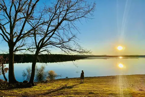 Una persona contempla el atardecer en la playa de Soria, con árboles desnudos y el sol reflejado en el agua serena.