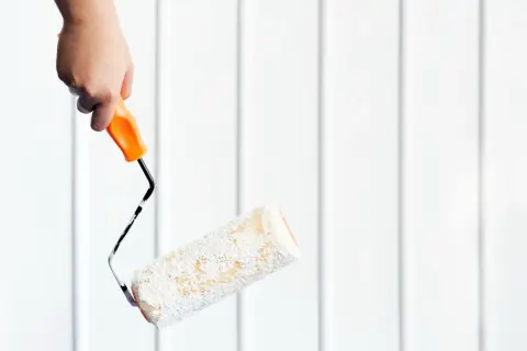 En hand håller en färgroller redo för målning utomhus mot en vit panelvägg.