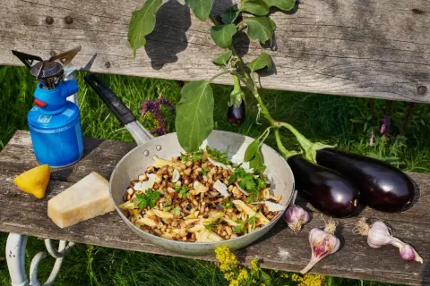 Pasta melanzane mit frischen Auberginen, Knoblauch und Parmesan. Ein Campingkocher und Zitrone deuten auf ein Outdoor-Mahl hin.