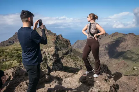 Un hombre fotografía el espectacular **barranco de Masca** mientras una mujer posa con las montañas y el mar de fondo.