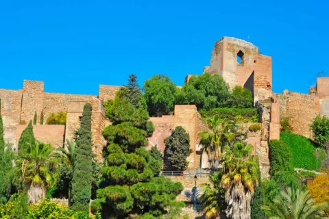 La Alcazaba de Málaga, fortaleza árabe con murallas de ladrillo y exuberante vegetación, se alza majestuosa bajo un cielo azul intenso.