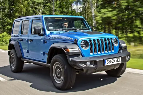Niebieski Jeep Wrangler 4xe Rubicon z kierowcą za kierownicą jedzie po drodze wśród drzew.