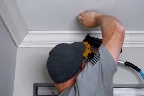 A man is performing crown molding installation, using a nail gun to secure the decorative trim to the ceiling.