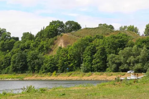 Zielona skarpa nad rzeką, na szczycie której widać ścieżki i ogrodzenie. To malownicza góra zamkowa w Drohiczynie.