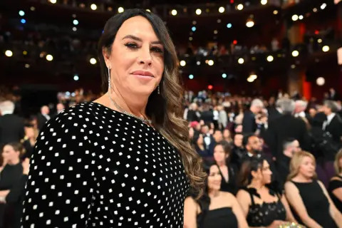 Karla Sofía Gascón, con un vestido negro de lunares blancos, sonríe en un evento con público al fondo.