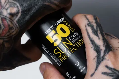 Hände mit Tattoos halten eine Sonnencreme-Spraydose. Die Dose schützt die Haut und das Tattoo vor der Sonne.