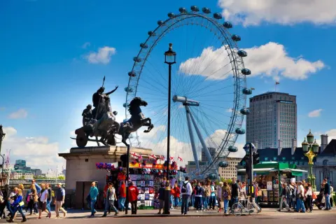 Fakta om London Eye: ett pariserhjul med gondoler, en staty av en häst och vagn, och en folkmassa på en solig dag.