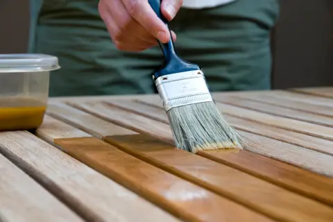 Application d'une lasure acrylique ou glycéro sur des lattes de bois. La brosse dépose le produit, transformant le bois clair en une teinte plus foncée et riche.