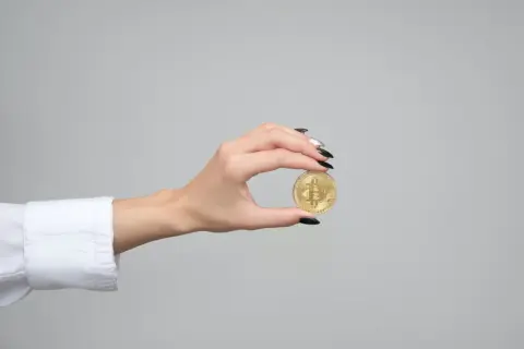 Dłoń z czarnym lakierem trzyma złotą monetę z symbolem Bitcoina. Zastanawiasz się, ile kosztuje złota moneta?
