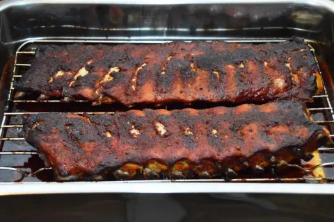 Zwei Rippchen-Stränge, dunkelbraun und saftig, ruhen auf einem Rost in einer Bratpfanne. Sie sind perfekt gegart, wie lange Spareribs im Ofen sein sollten.