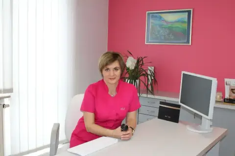 Lekarka w różowym uniformie siedzi przy biurku w gabinecie estetycznym.