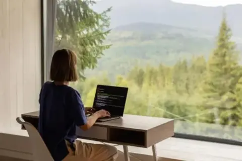 Persona trabajando en su portátil con vistas a un bosque y montañas. El estilo de vida de viajar trabajando es posible.