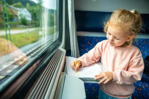 Mała dziewczynka rysuje kredką na kartce papieru, siedząc przy oknie w pociągu. Słońce oświetla jej twarz, tworząc magiczny pociąg słoneczny.