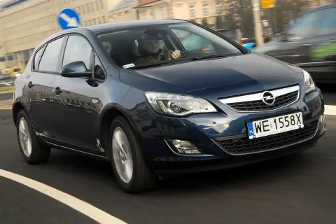Ciemnoniebieski Opel Astra J 1.7 CDTI porusza się po miejskiej drodze. Kierowca w czapce prowadzi auto.