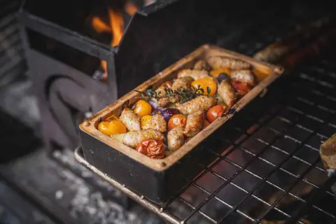 Salsiccia mit Gemüse in einer Auflaufform auf dem Grill, Flammen im Hintergrund.