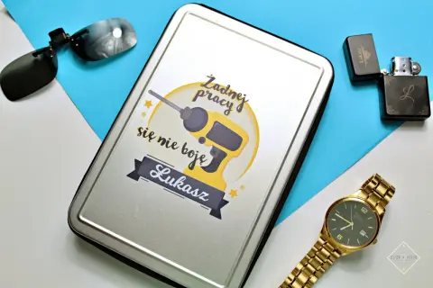 Prezent na dzień chłopaka dla Łukasza: metalowe pudełko z wiertarką i napisem "Żadnej pracy się nie boję", obok okulary, zapalniczka i złoty zegarek.