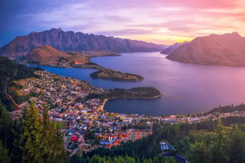 Vacker solnedgång över Queenstown, Nya Zeeland. Staden ligger vid en sjö, omgiven av berg och skog.