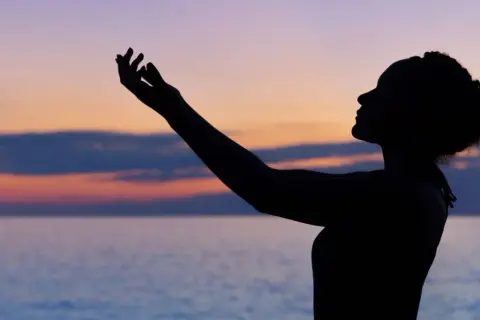 Silhouette einer Frau mit erhobenen Armen vor einem farbenfrohen Himmel und Meer. Sie scheint einen **chant deutsch** zu singen.