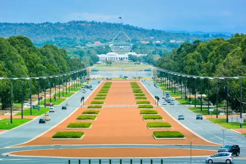 Widok na Parliament House, symbol tego, co jest stolicą Australii. Długa aleja z drzewami i samochodami prowadzi do budynku.
