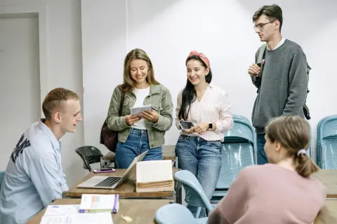 Grupa młodych ludzi dyskutuje o tym, ile trwa liceum dla dorosłych, przeglądając notatki i książki przy drewnianym stole.