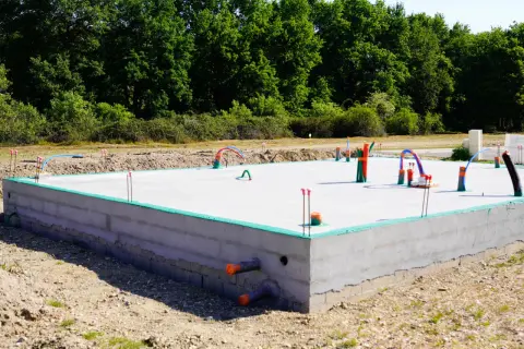 Płyta fundamentowa koszt budowy domu. Betonowa konstrukcja z instalacjami hydraulicznymi i elektrycznymi, otoczona zieloną izolacją i ziemią.
