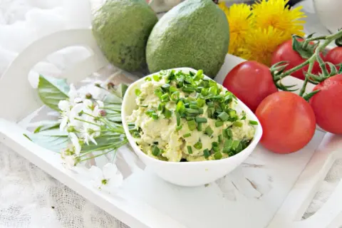 Pyszna pasta z awokado, jajkiem i szczypiorkiem, podana z pomidorkami i kwiatami na białej tacy.