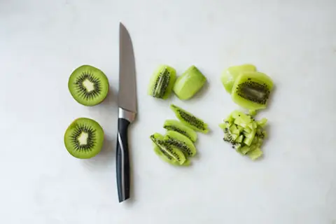 Ein Messer liegt neben zwei halbierten Kiwis. Daneben sind die Früchte in Scheiben und Würfel geschnitten, bereit zum kiwi schneiden.