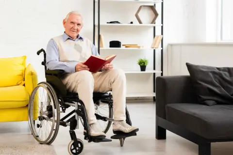 Anziano sorridente in carrozzina, circondato da ausili medici e arredi moderni, legge un libro.