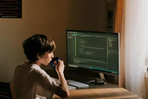 Un jeune garçon boit dans une tasse bleue devant un écran d'ordinateur affichant du code. Il est un développeur indépendant en herbe.
