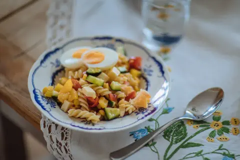 En färgglad pastasallad med ägg, grönsaker och ost, perfekt som pastasallad barn. En sked och ett glas vatten står bredvid.