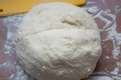 Surowe ciasto na pizzę na drewnianym blacie, posypane mąką. Obok żółta łopatka.