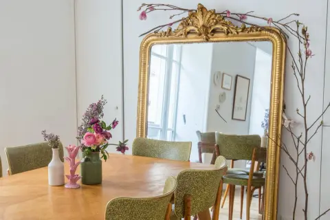 Un grand miroir au cadre doré orné de fleurs et de branches. Une table en bois est dressée avec des vases fleuris, prête à repeindre son cadre doré.