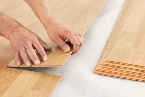 Hands installing laminate flooring over a subfloor. A stack of laminate planks is visible on the right. This shows how you can put laminate flooring over vinyl.