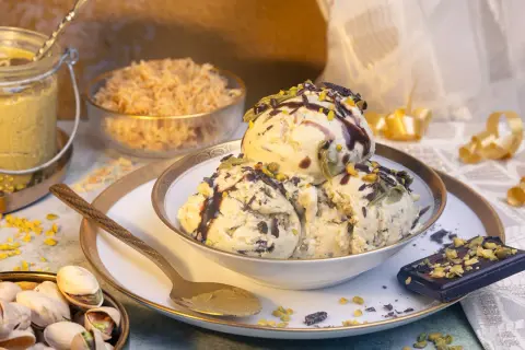 Schalen mit cremigem Pistazieneis, garniert mit Schokosauce und gehackten Pistazien. Daneben ein Stück dunkle Schokolade und eine Schale mit ganzen Pistazien.