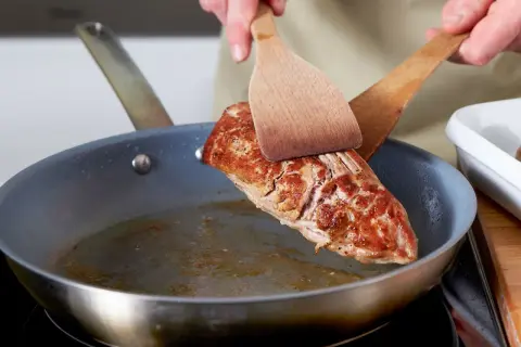 Ein saftiges Schweinefilet braten in der Pfanne. Mit zwei Holzspateln wird das Fleisch gewendet, um es von allen Seiten perfekt zu garen.