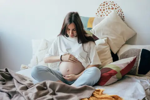 Kobieta w ciąży, otoczona poduszkami, czule dotyka brzucha. Czas oczekiwania na dziecko, być może zmagająca się z wielowodziem, ale pełna spokoju.